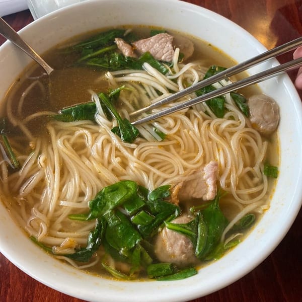 Close-up of a bowl of Thai noodle soup with meat, spinach, and bean sprouts, with chopsticks.